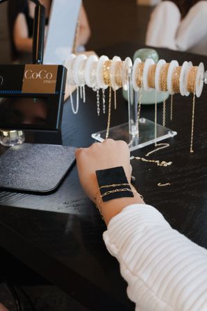 Wrist wearing delicate gold chain bracelets being tested against a display rack of gold and silver chains on a dark wood boutique table.