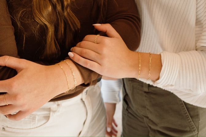 Close-up of two wrists wearing delicate layered gold chain bracelets with neutral tops and casual pants — minimalist jewelry style.
