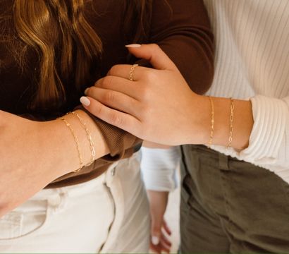 Close-up of two wrists wearing delicate layered gold chain bracelets with neutral tops and casual pants — minimalist jewelry style.