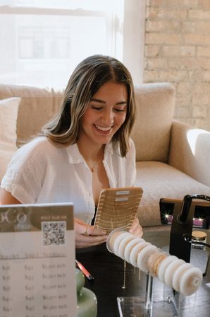 Smiling woman in a sunlit brick-walled living room joyfully browsing gold necklaces and bracelet display