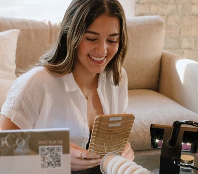 Smiling woman in a sunlit brick-walled living room joyfully browsing gold necklaces and bracelet display