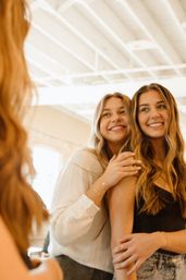 Two smiling young women with long wavy hair hug and look into a mirror in a bright sunlit studio — casual outfits and warm, candid friendship moment.