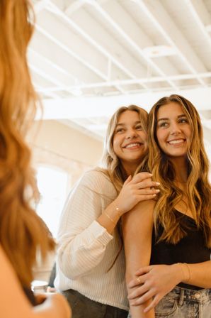 Two smiling young women with long wavy hair hug and look into a mirror in a bright sunlit studio — casual outfits and warm, candid friendship moment.