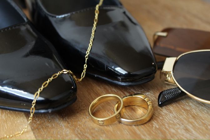 Close-up of glossy black patent shoes with a delicate gold chain, two gold rings and aviator sunglasses arranged on a wooden table