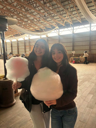 Two smiling friends holding oversized cotton candy inside a rustic wooden‑beam barn event space with draped fabric and vendor tables