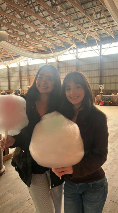Two smiling friends holding oversized cotton candy inside a rustic wooden‑beam barn event space with draped fabric and vendor tables