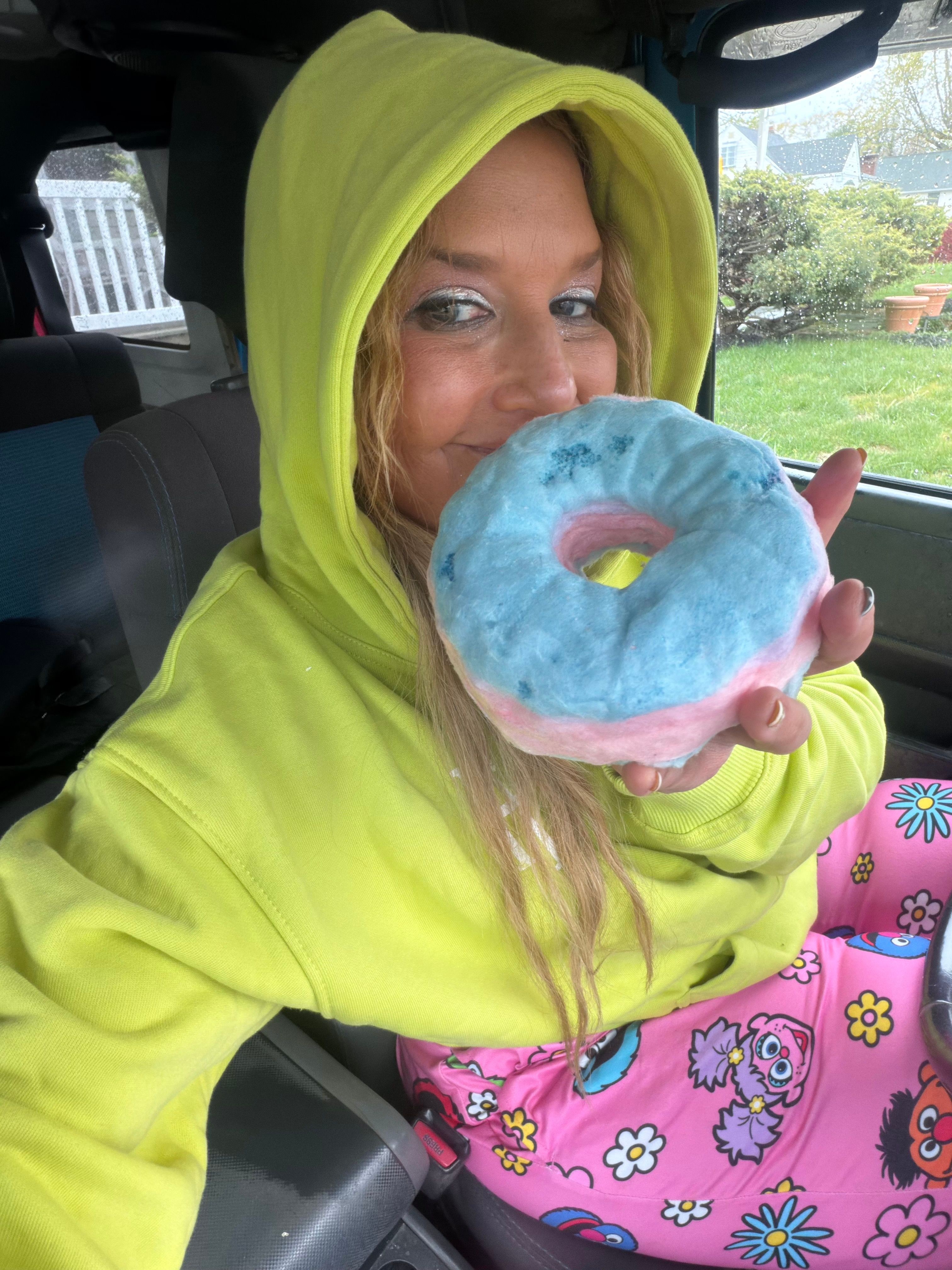 Person in a neon yellow hoodie in a parked car, smiling and holding a blue-and-pink frosted donut near their face; raindrops on the window and bright cartoon pajama pants add playful color.