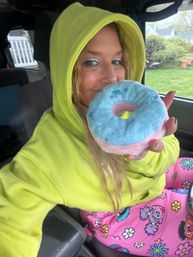 Person in a neon yellow hoodie in a parked car, smiling and holding a blue-and-pink frosted donut near their face; raindrops on the window and bright cartoon pajama pants add playful color.