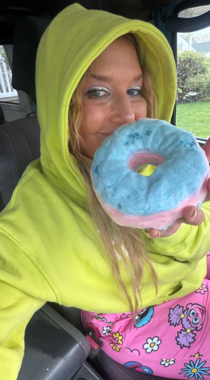 Person in a neon yellow hoodie in a parked car, smiling and holding a blue-and-pink frosted donut near their face; raindrops on the window and bright cartoon pajama pants add playful color.