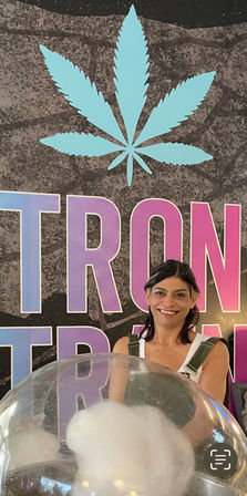 Smiling vendor making cotton candy beneath a large turquoise cannabis leaf logo and bold pink‑purple lettering on a textured wall.