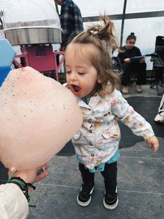 Toddler with a playful topknot leaning in to bite a giant pink cotton candy at a covered outdoor fair, cotton candy machine visible in the background