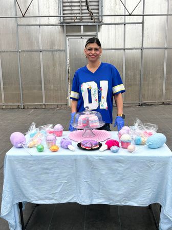Smiling vendor in a blue jersey at a greenhouse market stall selling colorful pastel bath bombs, cotton-candy soaps and layered soap slices displayed under a glass cloche.