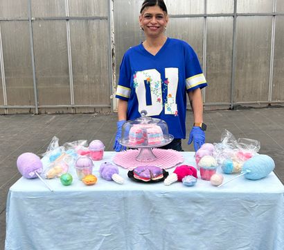 Smiling vendor in a blue jersey at a greenhouse market stall selling colorful pastel bath bombs, cotton-candy soaps and layered soap slices displayed under a glass cloche.
