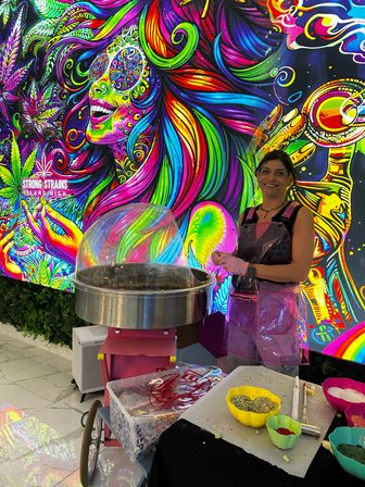 Smiling cotton candy vendor in a pink apron making treats at an indoor kiosk against a giant neon psychedelic mural with swirling rainbow hair, leafy motifs and colorful bowls of toppings.