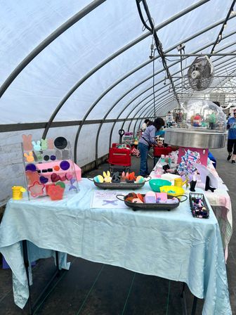 Greenhouse market stall in a curved hoop-house displaying colorful handmade soaps, bath bombs and treats on tables while shoppers browse near a cotton-candy machine and hanging fan.
