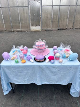 Pastel cotton-candy themed bath bombs, novelty soaps and treats arranged on a light blue tablecloth at a greenhouse vendor table.