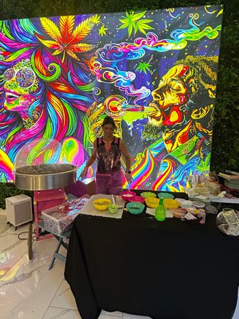 Vibrant neon psychedelic mural of colorful faces, swirling rainbow smoke and cannabis leaf motifs behind a vendor making cotton candy at a lively festival booth with bowls of bright sugar mixes on the table.