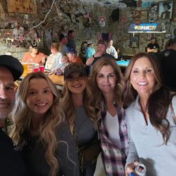 Friends taking a selfie inside a cozy small-town bar with dollar bills covering the walls, a pool table, wooden bar and neon Bud Light sign