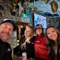 Group selfie of four people at a lively rustic bar with walls and ceiling covered in dollar bills and memorabilia, a TV and hanging hats above, a Modelo can on the counter and one person playfully giving the middle finger.