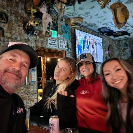Group selfie of four people at a lively rustic bar with walls and ceiling covered in dollar bills and memorabilia, a TV and hanging hats above, a Modelo can on the counter and one person playfully giving the middle finger.