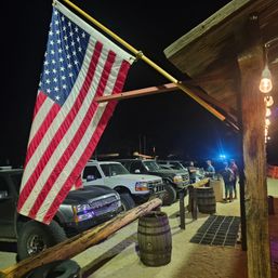 Large American flag hanging from a rustic wooden porch above a sandy nighttime parking area with lined-up pickup trucks, wooden barrels, and people chatting under string lights.