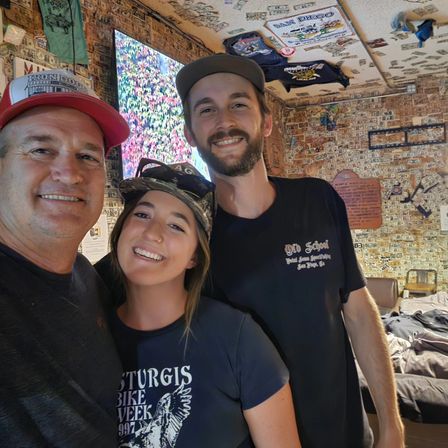 Three smiling people taking a selfie in a quirky San Diego bar plastered with dollar bills and hanging T-shirts