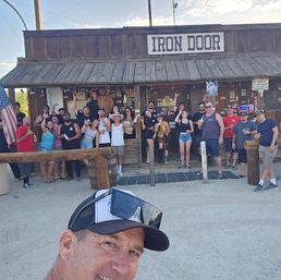 Selfie of a smiling man with sunglasses in front of a rustic wooden desert bar where a large group poses on the porch holding drinks, an American flag at left and a neon open sign visible under a sunny sky.