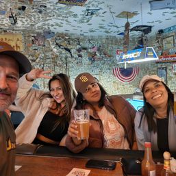Four friends smiling at a lively bar, one holding a beer, pool table and neon sign behind them, walls and ceiling plastered with dollar bills and an American flag bunting.