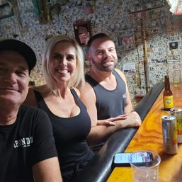 Three adults smiling for a selfie at a lively dive bar with walls covered in dollar bills, leaning on a wooden counter with beer bottles, cans and a smartphone.