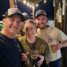 Three smiling friends posing with beer and wine glasses at a lively outdoor bar patio under hanging string lights at night