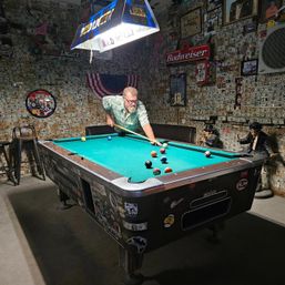 Person leaning over a teal billiards table taking a shot in a dim, Americana-themed bar with walls covered in dollar bills, a Budweiser sign, hanging beer lamp, and vintage decor.