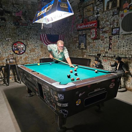 Person leaning over a teal billiards table taking a shot in a dim, Americana-themed bar with walls covered in dollar bills, a Budweiser sign, hanging beer lamp, and vintage decor.
