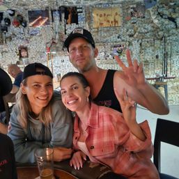 Three friends smiling and posing inside a lively bar with walls and ceiling covered in dollar bills and memorabilia, a beer glass on the table.