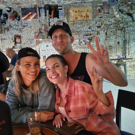 Three friends smiling and posing inside a lively bar with walls and ceiling covered in dollar bills and memorabilia, a beer glass on the table.