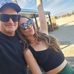 Two people wearing sunglasses take a cheerful selfie at a sunny desert spot with sandy ground, a small wooden shade structure with string lights, Adirondack chairs, and distant mountains on the horizon.