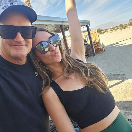 Two people wearing sunglasses take a cheerful selfie at a sunny desert spot with sandy ground, a small wooden shade structure with string lights, Adirondack chairs, and distant mountains on the horizon.