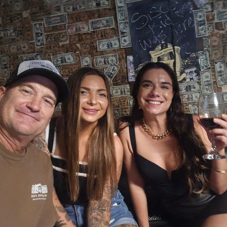Three friends smiling for a selfie in a lively bar with a wall covered in dollar bills; woman on the right raises a glass of red wine for a casual night out.