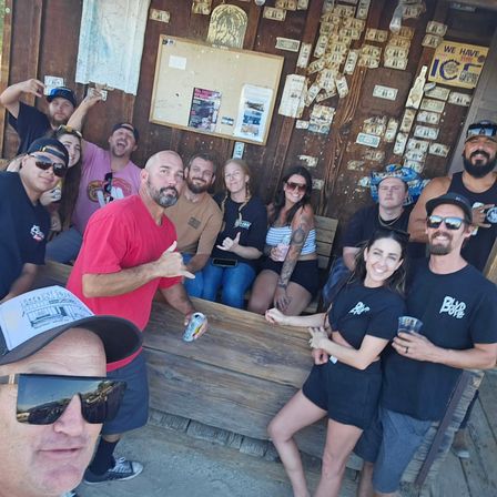 Selfie-style photo of a lively group of friends at a rustic outdoor wooden bar/shack plastered with dollar bills, smiling, holding drinks and making hand signs on a sunny day — casual summer attire and visible tattoos.