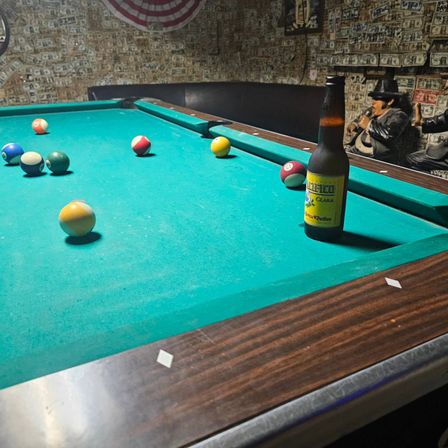 Green-felt pool table with scattered billiard balls and a beer bottle near the corner pocket in a dim bar lined with dollar-bill-covered walls and vintage decor.