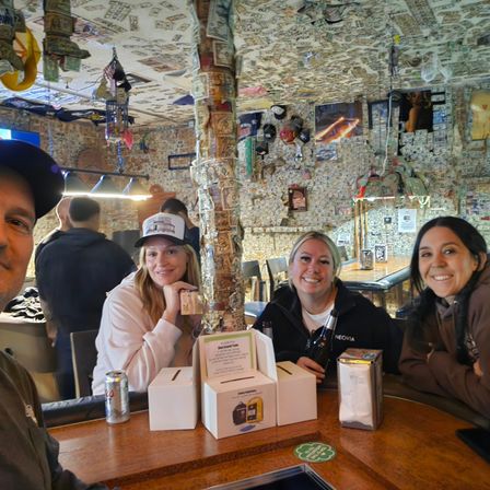 Four friends smiling at a lively dollar-bill-covered bar interior, wooden counter with drinks, napkin dispenser and donation boxes, walls and ceiling plastered with currency and memorabilia