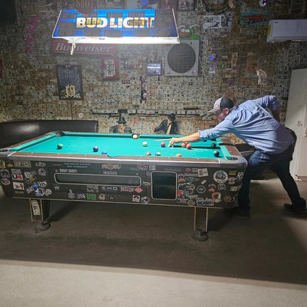 Person lining up a shot on a sticker-covered pool table beneath a hanging beer sign in a dim bar with walls plastered in dollar bills.