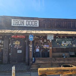 Sunlit rustic Old West–style saloon facade with a large painted sign, shaded wooden porch plastered with stickers and signs, a person standing in a coffin-shaped doorway, a handicapped parking sign, and a memorial banner.