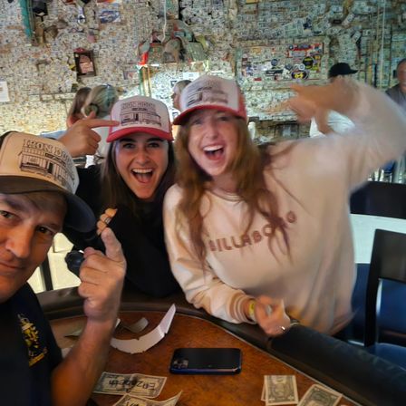 Three cheering friends wearing matching trucker hats in a lively bar with walls covered in dollar bills, posing over a table strewn with cash.