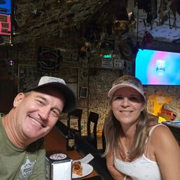 Smiling couple in caps at a lively dive bar with walls and ceiling plastered with dollar bills and quirky signs, sharing a slice of pizza and a beer at the bar counter with a TV in the background.