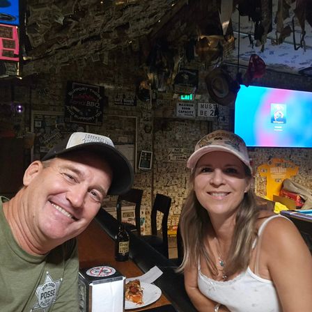 Smiling couple in caps at a lively dive bar with walls and ceiling plastered with dollar bills and quirky signs, sharing a slice of pizza and a beer at the bar counter with a TV in the background.
