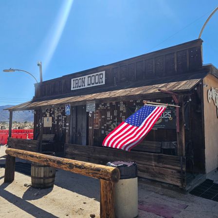 Sunny desert scene with a rustic wooden western storefront and porch, American flag waving, mountains and clear blue sky in background