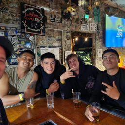 Five friends smiling at a wooden bar counter, holding beers and making hand signs, with walls covered in dollar bills, stickers and neon signs — lively night out