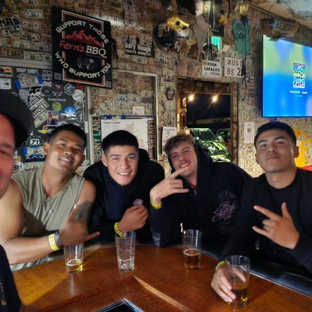 Five friends smiling at a wooden bar counter, holding beers and making hand signs, with walls covered in dollar bills, stickers and neon signs — lively night out