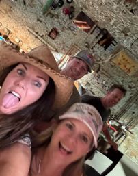 Playful group selfie of four friends in a rustic Western-themed room — woman in a cowboy hat sticking her tongue out, woman in a pink cap smiling, two men laughing in the background.