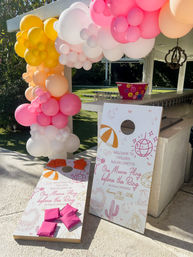 Poolside Palm Springs bachelorette setup with two custom cornhole boards, pink and orange beanbags and a playful pink, peach, yellow and white balloon garland draped over a bar.